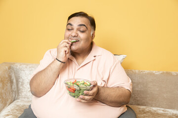 Happy Overweight fat indian man eating healthy vegetable salad food at home. Plus size guy enjoy meal to lose weight. Dieting and Obesity concept.