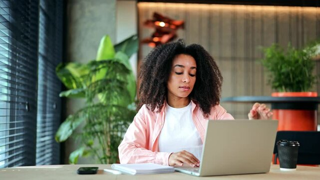 Focused African American businesswoman, dressed in a pink shirt, opens and types on her laptop at a modern desk. Gitl is engaged in productive remote work in a contemporary office setting.