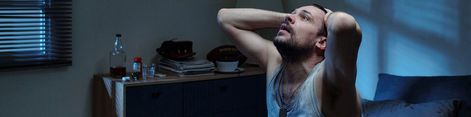 Fototapeta premium Caucasian man sitting on bed holding head in hands looking up appearing distressed in dimly lit bedroom suggesting emotional struggle related to family problems