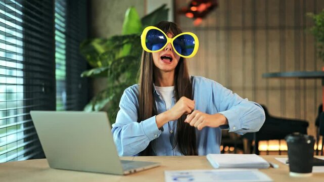Cheerful businesswoman in light blue shirt and oversized yellow glasses dances playfully at office desk. She expresses pure joy and happiness, enjoying a fun moment during work in a modern workspace.