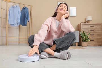 Young woman in headphones turning on robot vacuum cleaner at home
