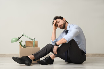 Upset fired young man with box of personal stuff sitting near light wall