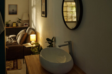 Empty bathroom with modern sink and round mirror in foreground, bedroom with unmade bed and warm...