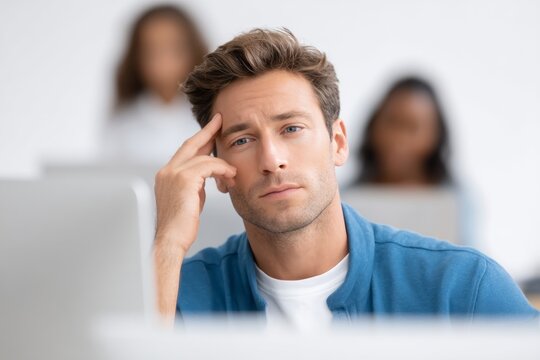 Thoughtful young caucasian male adult in blue sweater working at desk with colleagues