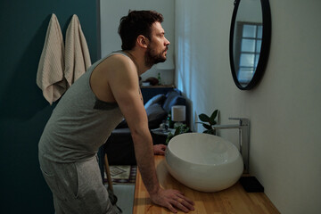 Caucasian young adult man leaning over bathroom sink staring into mirror with serious expression,...