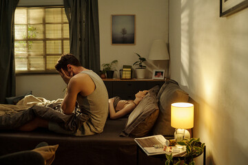 Young adult man sitting on bed covering face with hands while woman lying beside him sleeping,...