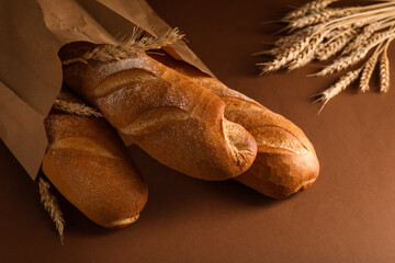 Paper bag with fresh baguettes and wheat on brown background, closeup