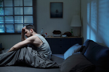 Caucasian man sitting on bed holding head in hand appearing distressed in dimly lit bedroom...