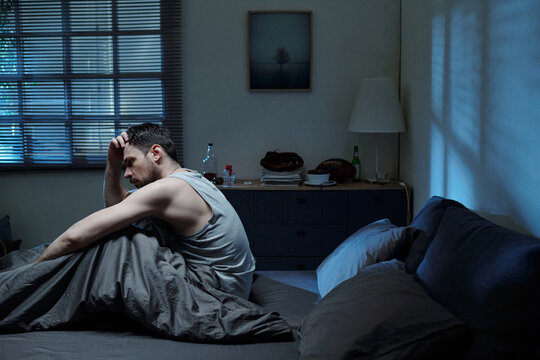Caucasian man sitting on bed holding head in hand appearing distressed in dimly lit bedroom suggesting family problems, looking away from camera with tense posture - Powered by Adobe