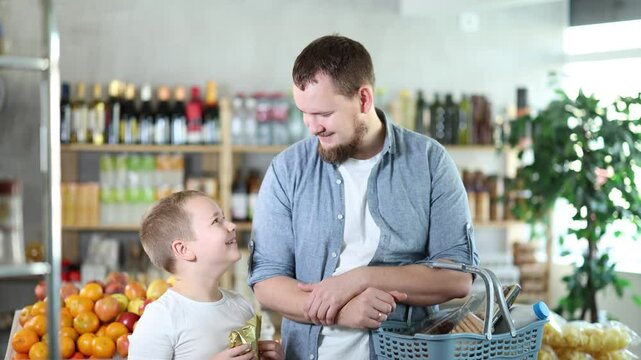 Visiting store, father and son is looking at something on shelf of showcase. Family holds heavy basket of groceries and reaches for shelf, trying to see products in refrigerator window.