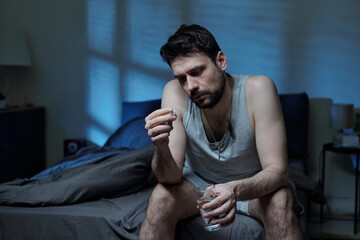Caucasian young adult man sitting on bed holding pill in one hand and glass of water in other hand,...