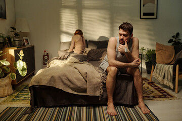 Man sitting on bed looking away with thoughtful expression while woman sitting on bed with back...
