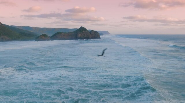 Aerial view of waves crashing against the rocky coastline of Bethells Beach, New Zealand. The rugged landscape and powerful ocean create a dramatic scene.