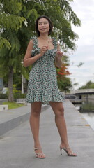 Asian woman holding her sunglasses stands in a floral dress and stylish high-heeled sandals,...
