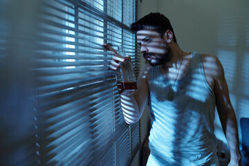 Caucasian man holding whiskey bottle standing by window blinds peering outside with tense...