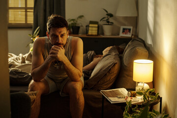 Caucasian young adult man sitting on bed with hands clasped, appearing deep in thought while woman...