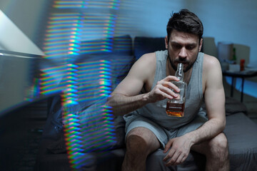 Caucasian young adult man sitting on bed drinking whiskey from bottle, appearing distressed and...