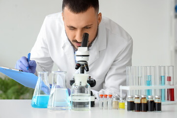 Male scientist with clipboard looking through microscope at table in laboratory