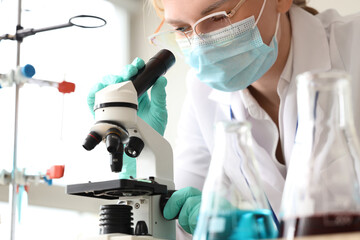 Female scientist with medical mask looking through microscope in laboratory, closeup