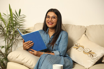 Pretty Young woman sitting on cozy couch sofa and relaxing in her living room reading book in...