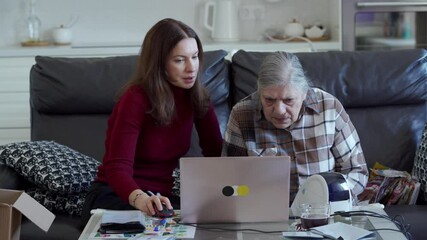 A woman assists her senior mother in managing their online store, using a laptop and handling shipping tasks on a table in their home's living room