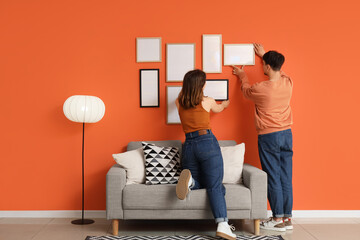 Young couple hanging blank frames on orange wall at home, back view