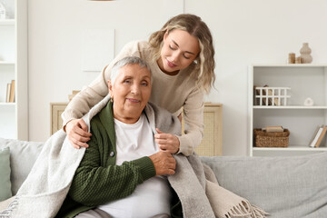 Young woman covering her grandmother with warm plaid on sofa at home