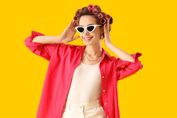 Young woman in sunglasses with hair curlers on yellow background
