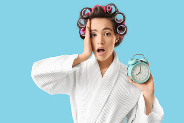 Shocked young woman in bathrobe with hair curlers and alarm clock on blue background