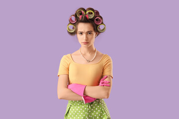 Upset young housewife with hair curlers on lilac background