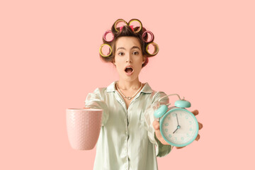 Shocked young woman in pajamas with hair curlers holding coffee cup and alarm clock on pink...