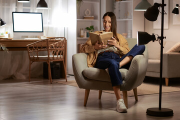 Beautiful woman reading book on armchair at home in evening