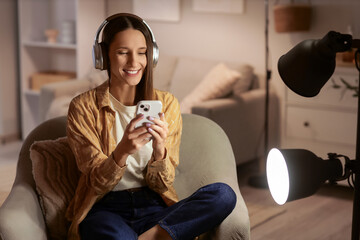 Beautiful woman with headphones using mobile phone on armchair at home in evening