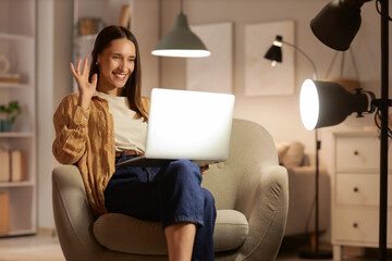 Beautiful woman with laptop video chatting on armchair at home in evening
