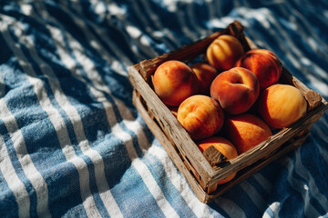 Juicy ripe peaches arranged in a wooden basket placed on a striped blue and white cloth in natural outdoor light, suggesting summer freshness.