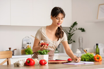 Young woman writing recipe in book while cooking pizza at table in kitchen
