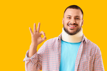 Handsome young man with neck splint showing OK gesture on yellow background