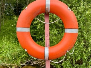 An orange and white lifebuoy hangs on the dock against a backdrop of blooming greenery by the...