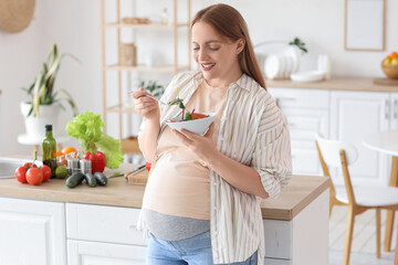 Young pregnant woman eating vegetable salad in kitchen