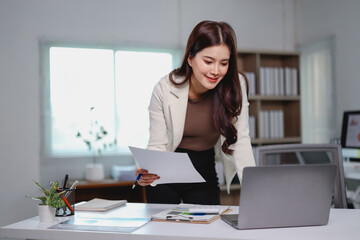 Young asian businesswoman holding documents and comparing information with laptop, working on...