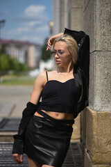 Fashionable beautiful girl in black blazer and black leather skirt posing on urban background.