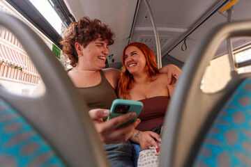 Two joyful female friends enjoying their bus trip, browsing social media on a smartphone and...