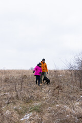 Naklejka premium Exploring the tranquil landscape on a cloudy day, a person in an orange jacket and another in a pink coat walk their dog through golden grasses and the soft earth path