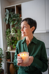 Calm thoughtful mature woman with scented candle relaxing in thoughts at home. Female meditation, aromatherapy benefit, wellness mental clarity, slow living hygge, interior fragrance decoration 