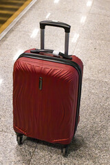 A red hard-shell suitcase with a telescopic handle standing on a polished granite floor at an airport terminal. Travel and luggage concept.