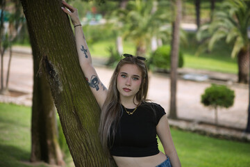 Portrait of a young woman, beautiful, blonde, green eyes, wearing a black t-shirt, looking at the camera sweetly and tenderly leaning against the trunk of a tree. Concept of tenderness, innocence.