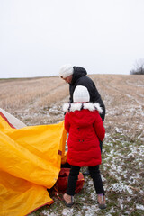 Preparing for a winter adventure as a child and adult set up a bright yellow tent in a snowy field under overcast skies