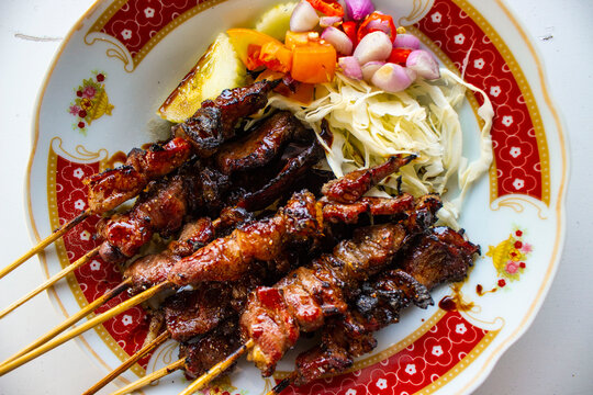 sate kambing or sate klatak or lamb satay or goat meat satay served with served with sliced ​​cabbage, ​​red onion, ​​chili, ​​tomatoes with soy sauce on plate. isolated white background -