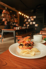 Close-up of gourmet sandwich bagel with shredded veggie mushrooms and chips served on white plate in cozy café setting with warm lighting and blurred background of diners and decor. Asunción, Paraguay
