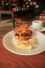 Close-up high angle shot of gourmet vegetarian sandwich bagel with mushrooms; chips served on white plate in cozy café setting. Coffee on table in background. Travel photography in Asunción, Paraguay